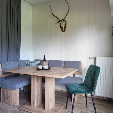 A cozy dining area with a wooden table and a bench. A deer antler hangs on the wall, and there is a green chair.