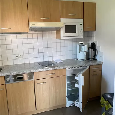 Eine moderne Küche mit Holzschränken und weißen Fliesen. Es gibt einen Kühlschrank, eine Mikrowelle und gängige Küchengeräte.