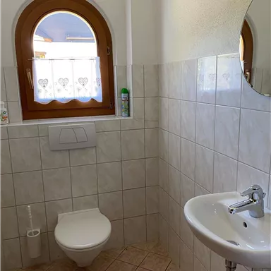 Ein Badezimmer mit keramischen Fliesen und einem ovalen Fenster. Es gibt ein WC und ein Handwaschbecken.