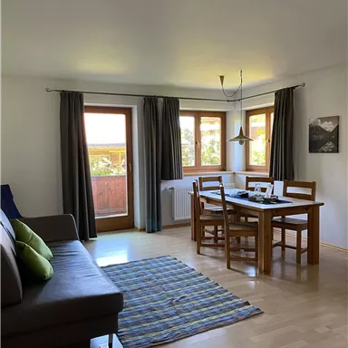 Ein gemütlicher Wohnraum mit einem Esstisch und einer Couch. Große Fenster sorgen für viel Tageslicht und einen Blick nach draußen.