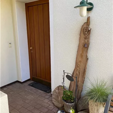 Eine einladende Haustür aus Holz mit einer Wandlampe daneben. Vor der Tür steht eine kleine Pflanzenanordnung und ein dekoratives Stück Holz.