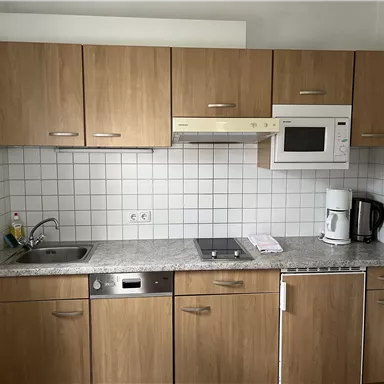 Eine moderne Küche mit Holzschränken, einem Spülbecken und einem Mikrowellenofen. Die Arbeitsfläche ist aus grauem Stein und es gibt verschiedene Küchengeräte.