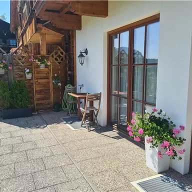 Ein einladender Eingangsbereich mit einer Terrasse, Pflanzen und einem kleinen Tisch. Das Gebäude im Hintergrund zeigt traditionelle Architektur.
