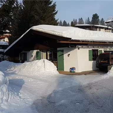 Ein gemütliches Chalet, umgeben von viel Schnee und Bäumen. Die Sonne scheint auf das Dach, das mit einer Schneeschicht bedeckt ist.