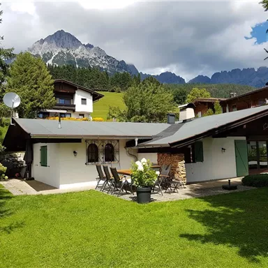 Ein idyllisches Haus in den Bergen, umgeben von grünem Rasen und Bäumen. Im Hintergrund sind majestätische Berge und ein teilweise bewölkter Himmel zu sehen.