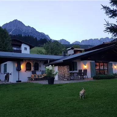 Ein charmantes Haus in den Bergen, umgeben von Wiesen und Bäumen. Im Vordergrund läuft ein kleiner Hund über den Rasen.