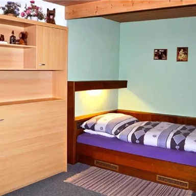 Ein schlicht eingerichtetes Schlafzimmer mit einem Einzelbett und einem Holzregal. Die Wände sind in sanften Farben gestrichen und es gibt einige persönliche Gegenstände.