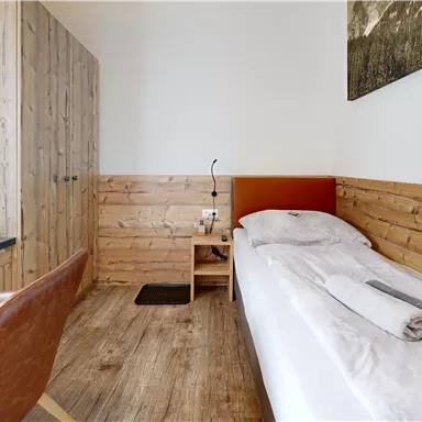 A simple room with wooden paneling and a single bed. There is a desk, a chair, and a window with curtains.