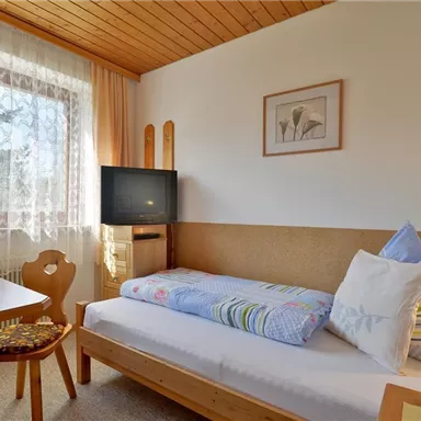 Ein gemütliches Zimmer mit einem Einzelbett und einem Tisch. Das Zimmer hat Holzdetails und großen Fenstern, die viel Licht hereinlassen.