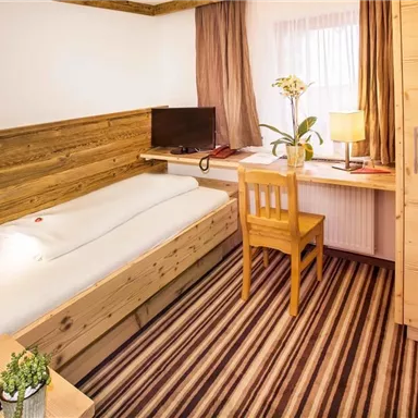 A cozy bedroom with wooden furniture and a single bed. There is a television at the workspace, and a desk with a lamp and a plant completes the decor.