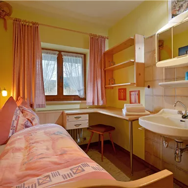 Ein gemütliches Zimmer mit einem Einzelbett und einem kleinen Tisch. Die Wände sind gelb gestrichen, und es gibt ein Fenster mit Blick nach draußen.