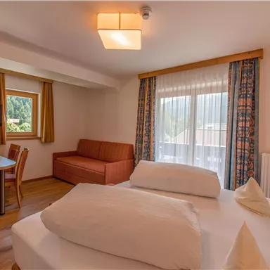 Ein gemütliches Zimmer mit einem Doppelbett und einer Sitzecke. Große Fenster sorgen für viel Tageslicht und einen schönen Ausblick.