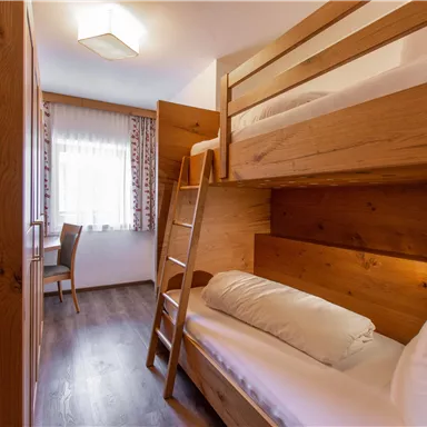 Ein gemütliches Zimmer mit einem Etagenbett aus Holz und einem einzelnen Bett. Ein Tisch und ein Stuhl ergänzen die Einrichtung, und das Fenster lässt viel Licht herein.