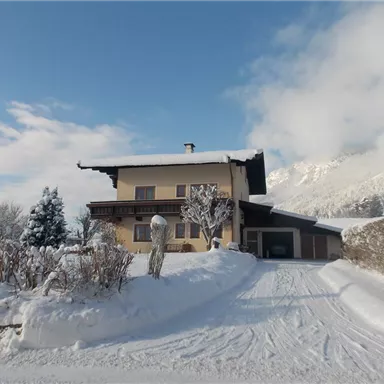 A cozy house in the snow with a snow-covered driveway. In the background, there are snow-covered mountains and a clear sky.