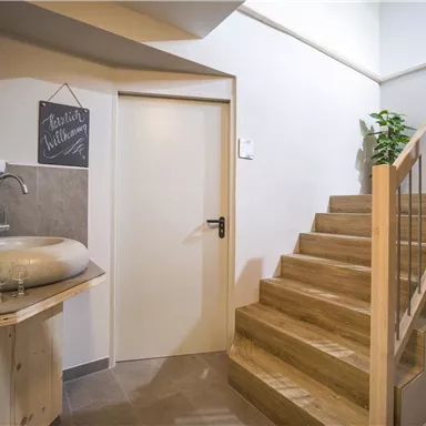 A modern entrance area with a wooden staircase and an elegant sink. A small board hangs on the wall and there are some plants for decoration.