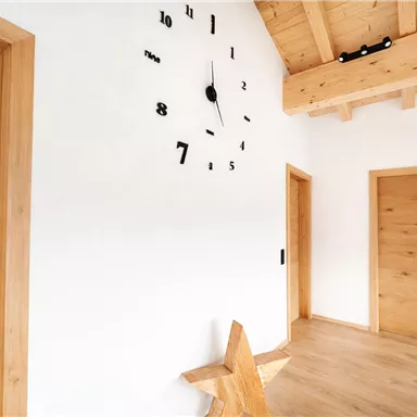 Ein heller Raum mit Holzdetails und einer großen Uhr an der Wand. Es gibt zwei Türen und eine dekorative Holzsternfigur auf dem Boden.