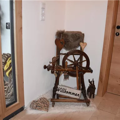 Eine traditionelle Spinning Wheel und ein Willkommen-Schild stehen in einer gemütlichen Ecke. Die Umgebung besteht aus Holz und zeichnet sich durch einen rustikalen Charme aus.