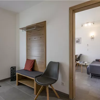 A modern entrance area with a wooden bench and a chair. The walls are light, and there is a view of an adjacent room.