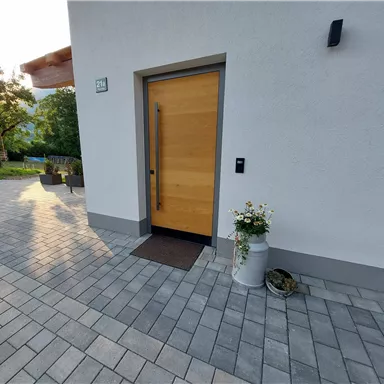 Eine moderne Eingangstür aus Holz mit einem matten Fußabtreter. Neben der Tür steht ein Blumentopf mit weißen Blumen.