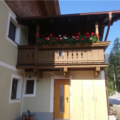 Ein schönes Haus mit einem Balkon, der mit roten Blumen geschmückt ist. Der Himmel ist klar und blau, was für eine sonnige Atmosphäre sorgt.