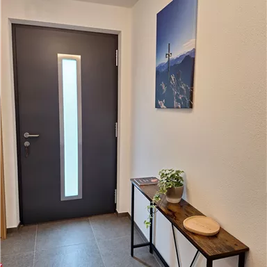 Ein modern gestalteter Eingangsbereich mit einer dunklen Tür und einem Wandbild von Bergen. Auf einem kleinen Tisch stehen eine Pflanze und ein Holzbrett.