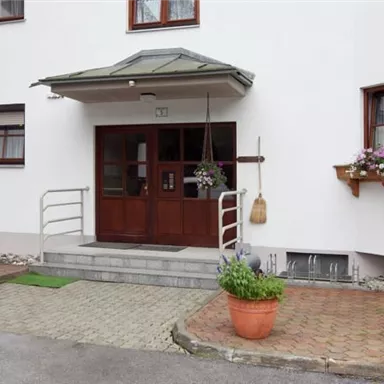 Ein heller Eingangsbereich eines Gebäudes mit einer Stuttür und Fenstern. Blühende Pflanzen stehen in Töpfen auf der Fensterbank.