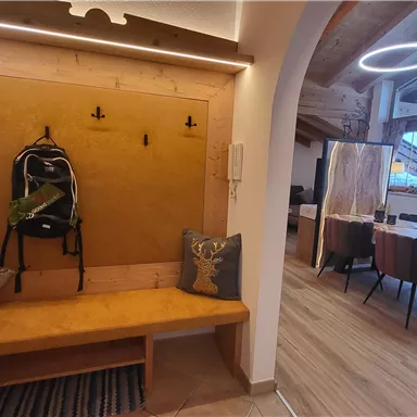 Ein moderner Eingangsbereich mit einer Holzbank, einem Rucksack und einem hängenden Regal. Im Hintergrund ist ein hell erleuchteter Essbereich zu sehen.