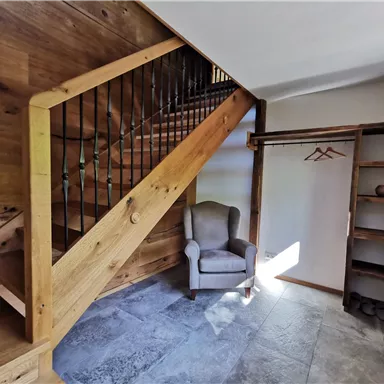 A bright entrance area with a wooden staircase and a cozy armchair. On the wall, there is an open shelf and some coat hooks.
