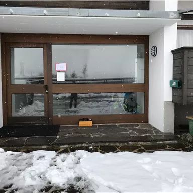 An entrance with large windows and a wooden porch. Outside, there is snow and it is lightly snowing.
