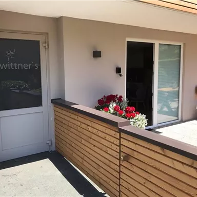 A modern entrance to a building with the inscription "wittner's".  
Flowers in colorful boxes adorn the area in front of the door.