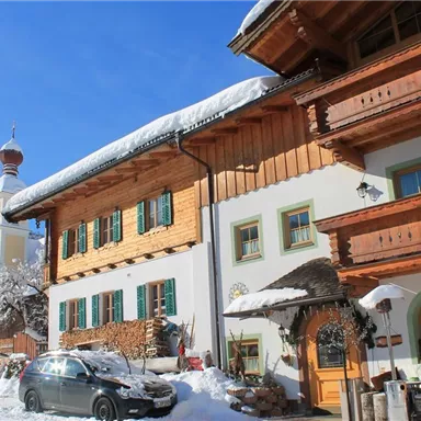 Ein malerisches Almhaus im Winter mit schneebedecktem Dach und bunten Fenstern. Im Hintergrund steht ein Kirchturm vor einer schönen Berglandschaft.