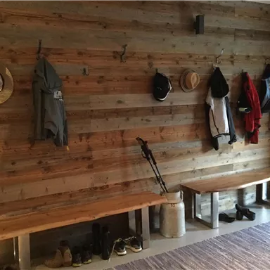 Eine rustikale Wand mit Holzverkleidung, an der Kleidung und Hüte hängen. Darunter befinden sich zwei Bänke und Schuhe.