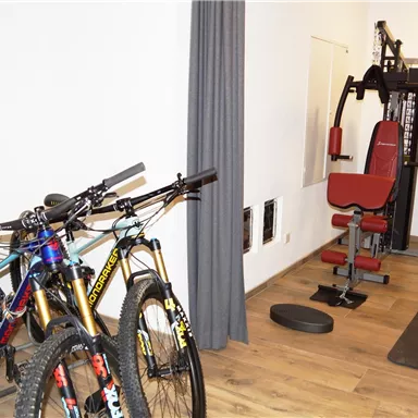 Ein Fitnessraum mit zwei Mountainbikes und einem Multifunktionsgerät. Der Raum hat einen Holzboden und graue Vorhänge.
