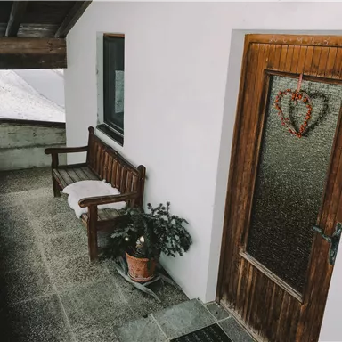 Eine gemütliche Veranda mit einer Holzbank und einem kleinen Topf mit Pflanze. Die Eingangstür hat ein herzförmiges Dekor.