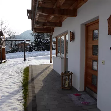 A cozy house in winter with snow on the ground. In the background, playground equipment and trees can be seen.