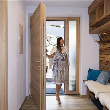 A woman is standing at the open door of a modern entrance area. The room is bright and decorated with wooden paneling.
