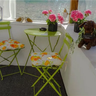 Ein sonniger Wintergarten mit einem kleinen grünen Tisch und zwei Stühlen. Blumen und eine dekorative Hundestatue verleihen der Szene einen einladenden Charme.