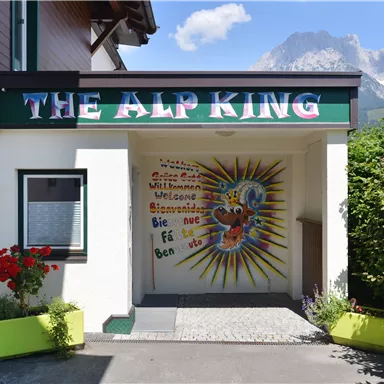 Ein Eingang zu einem Gebäude mit dem Schriftzug "The Alp King". Die Wand ist bunt gestaltet und zeigt eine freundliche Begrüßung.