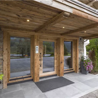 Eingang eines holzverkleideten Gebäudes mit großen Glasfenstern. Das Foyer ist von grünen Pflanzen und blühenden Blumen umgeben.