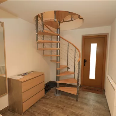 Ein heller Eingangsbereich mit einer eleganten Wendeltreppe aus Holz und Metall. Rechts befindet sich eine Tür mit Glasinnenteilen und eine moderne Kommode.