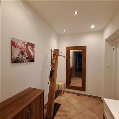 Ein heller Flur mit einer Holzkommode und einem Spiegel. An der Wand hängt ein gemaltes Bild und ein Garderobenständer steht in der Ecke.