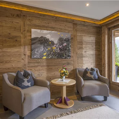 Ein gemütlicher Innenraum mit Holzverkleidung und zwei beigen Sesseln. In der Mitte steht ein runder Tisch mit einer Blumendekoration und einem großen Fenster, das auf eine grüne Landschaft blickt.