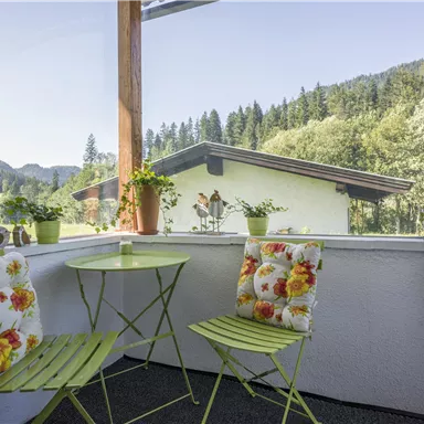 Ein gemütlicher Balkon mit grünen Stühlen und einem kleinen Tisch. Im Hintergrund sind Bäume und ein Haus in einer malerischen Landschaft zu sehen.