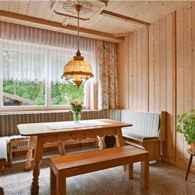 Ein gemütlicher Essbereich aus Holz mit einer hellen Lampe und einer großen Fensterfront. Im Raum stehen ein Tisch mit Bänken und ein grüner Pflanzenaufsteller.