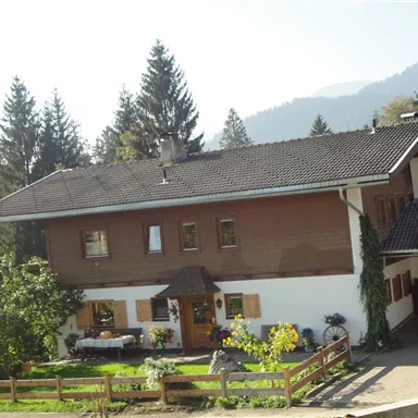 Ein gemütliches, traditionelles Haus umgeben von Bäumen und blühenden Pflanzen. Im Hintergrund sind sanfte Hügel zu sehen.