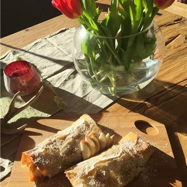 Two delicious apple strudels on a wooden board, surrounded by fresh tulips in a glass vase. The warm lighting gives the scene a cozy atmosphere.