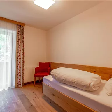 Ein schlichtes Zimmer mit einem Einzelbett und einem roten Stuhl. Große Fenster sorgen für viel Licht und einen Blick nach draußen.