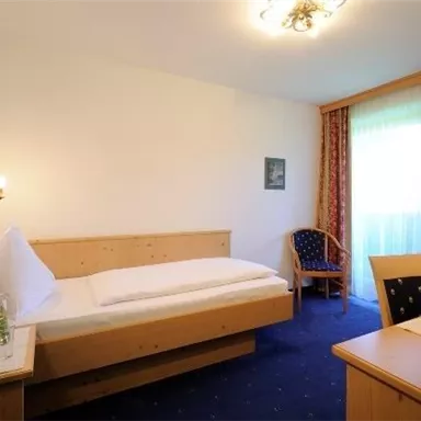 Ein einfaches und gemütliches Einzelzimmer mit Holzmöbeln. Es gibt ein Bett, einen Tisch und einen Stuhl sowie ein Fenster mit Vorhängen.