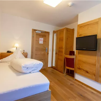 Ein einfaches Zimmer mit einem Einzelbett und Holzdetails. Es gibt einen Fernseher und einen kleinen Tisch mit einem Stuhl.