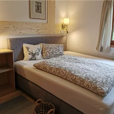 Ein gemütliches Schlafzimmer mit einem Doppelbett und einer hellen, freundlichen Atmosphäre. Neben dem Bett steht ein Holzregal und das Fenster lässt viel Tageslicht herein.